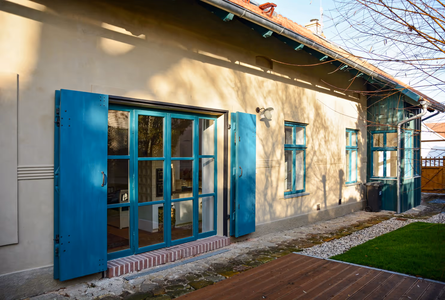 Exterior of a light beige house with blue window frames and shutters, a wooden deck, and a small green lawn.