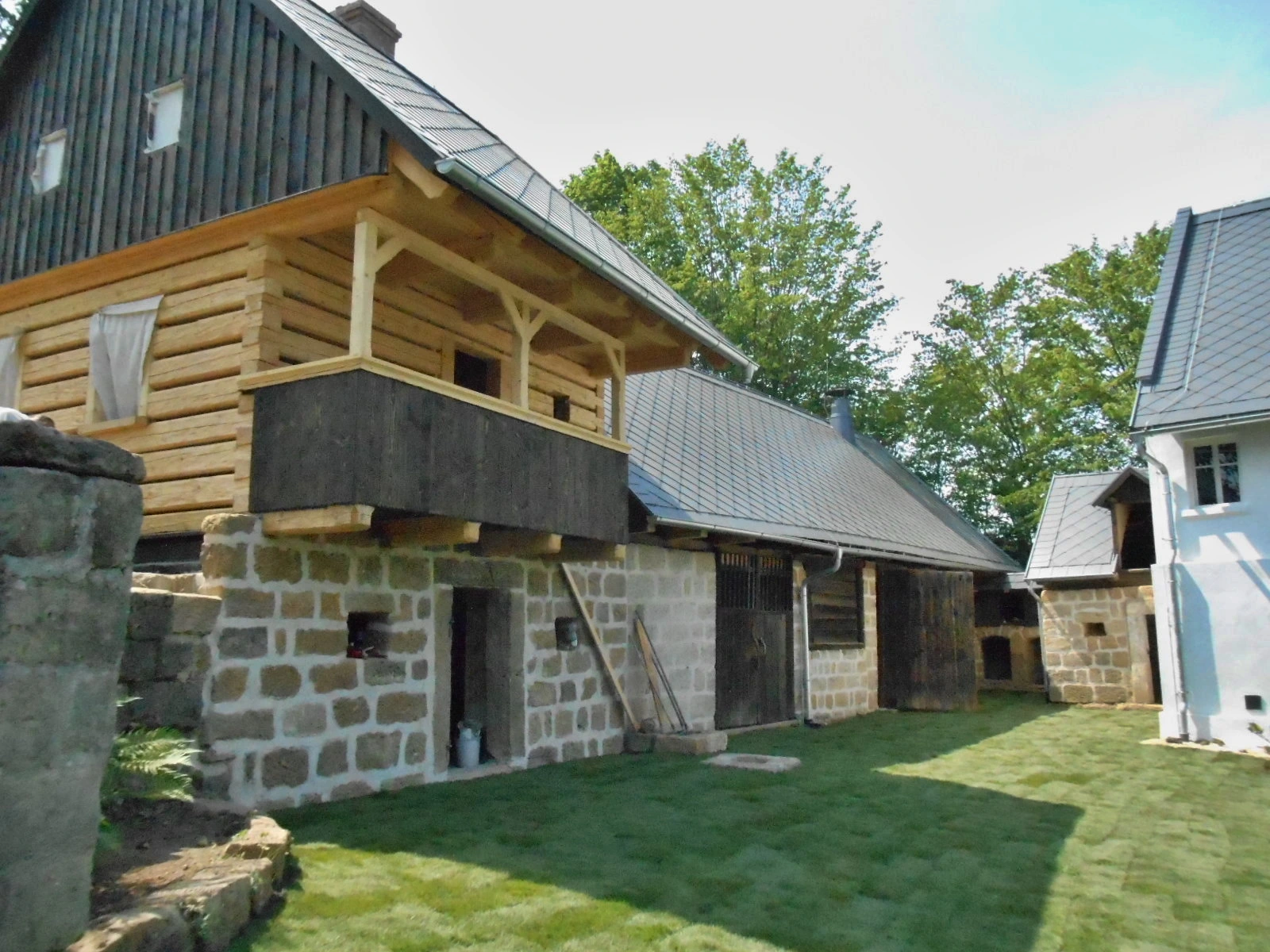 Traditional stone and wood house with a gabled roof, wooden balcony, and green lawn under a blue sky with trees in the background.