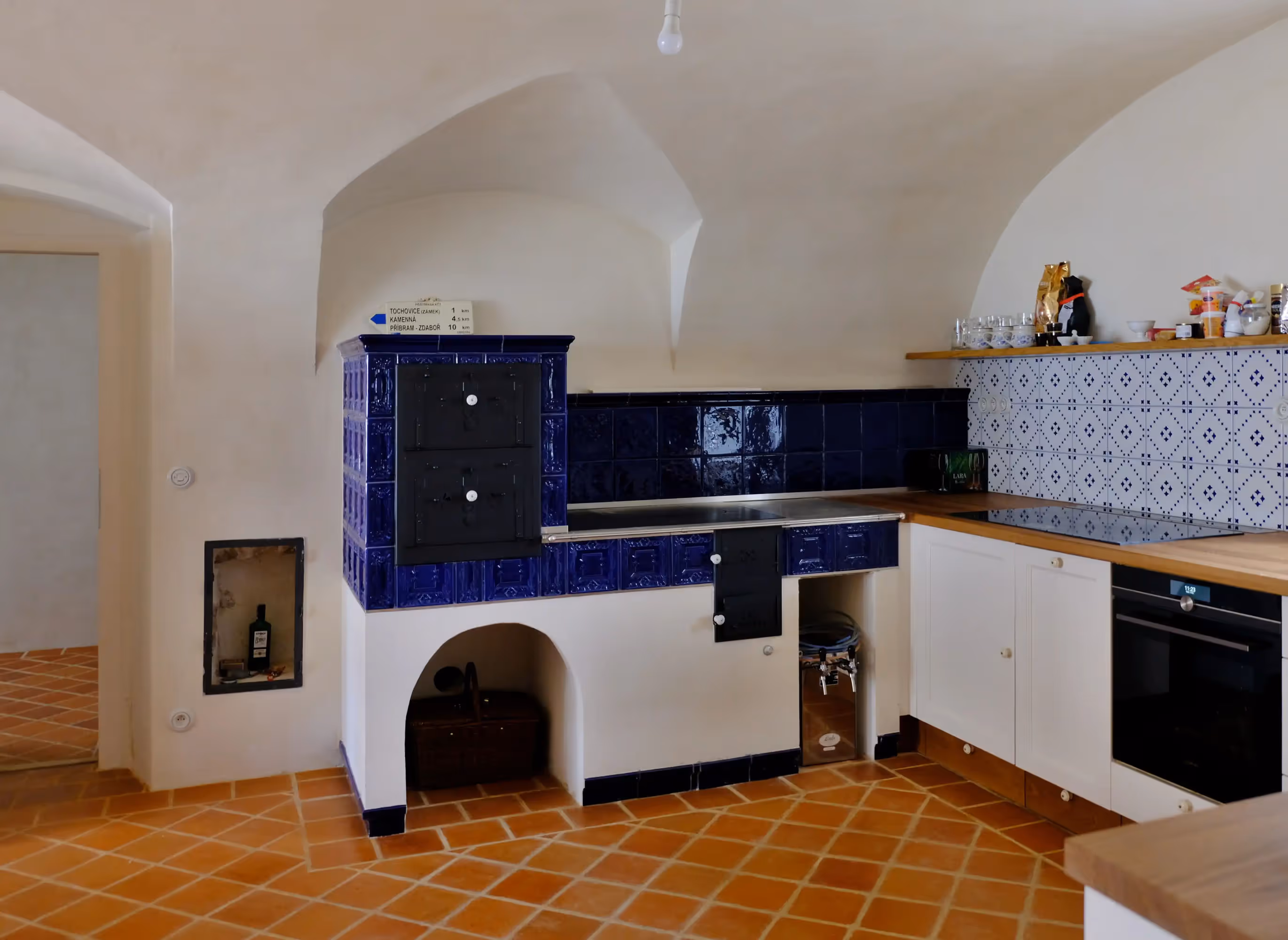 Rustic kitchen with terracotta tiled floor, white walls, blue tiled stove, wooden countertops, and white cabinetry.