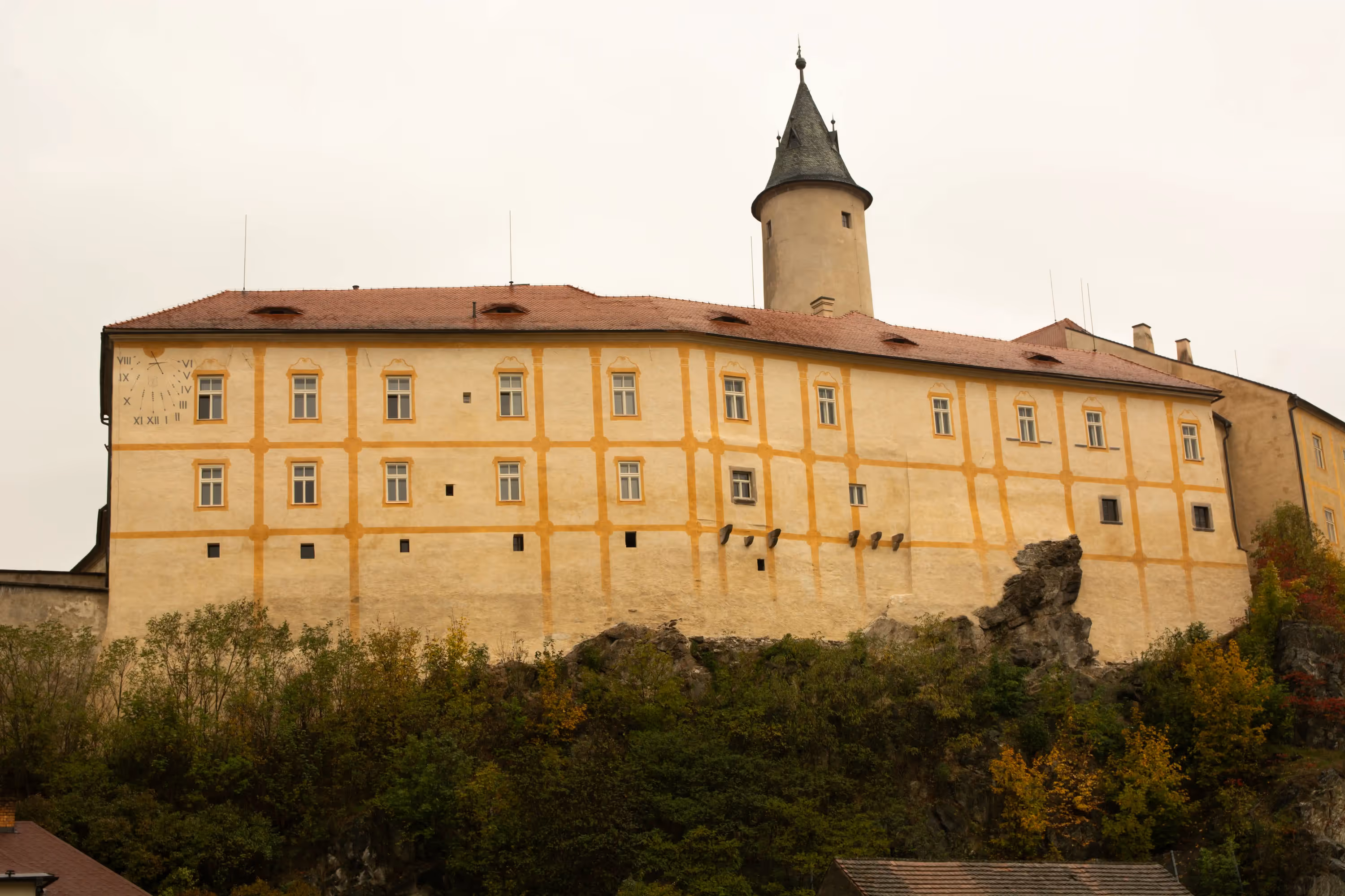 Historic beige castle with a red-tiled roof, a round tower with a cone-shaped roof, and Roman numeral clock on the wall, perched on a rocky hill covered with trees.