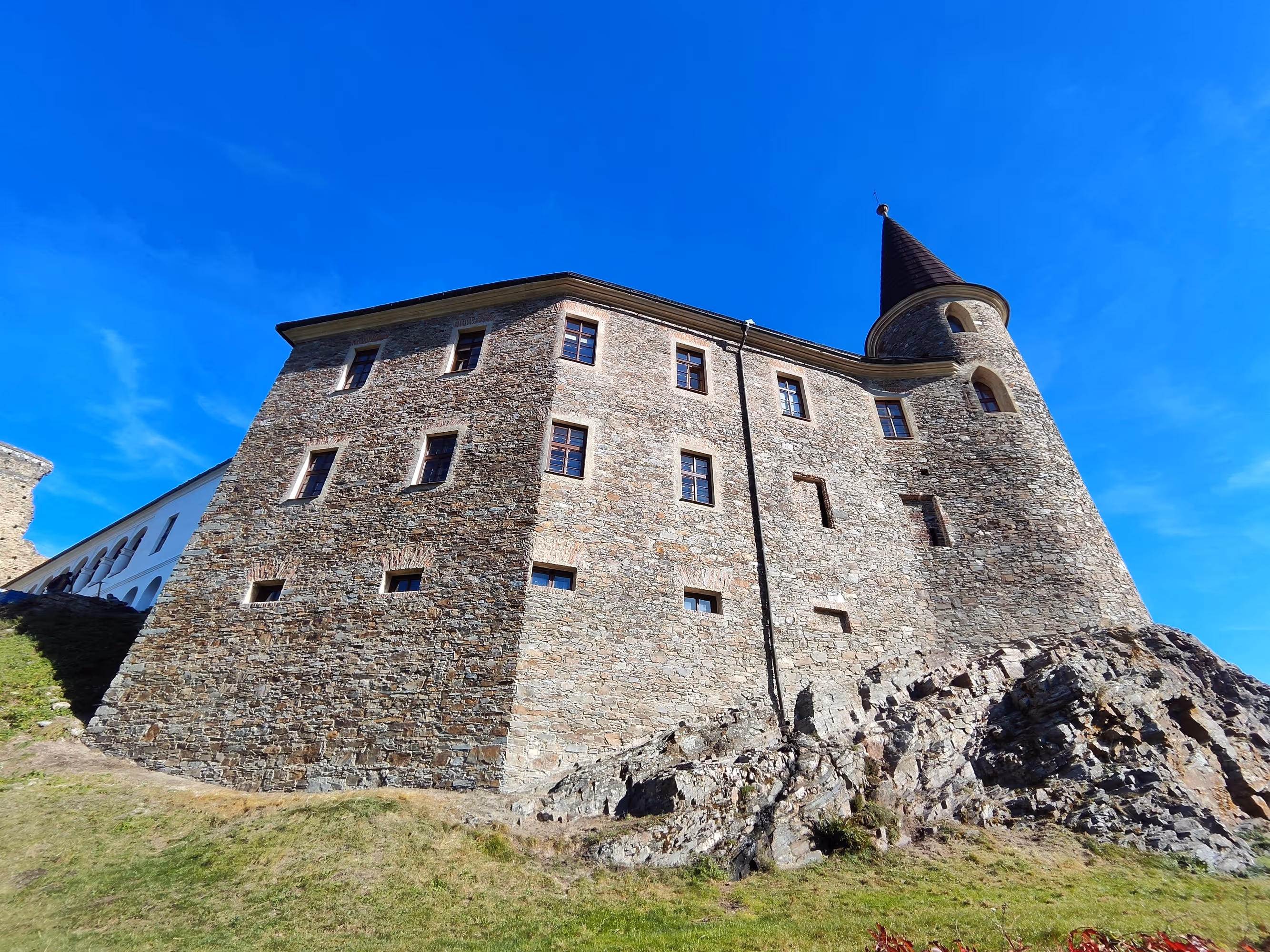 Stone castle with multiple windows and a conical tower on a rocky grassy hill under a clear blue sky.