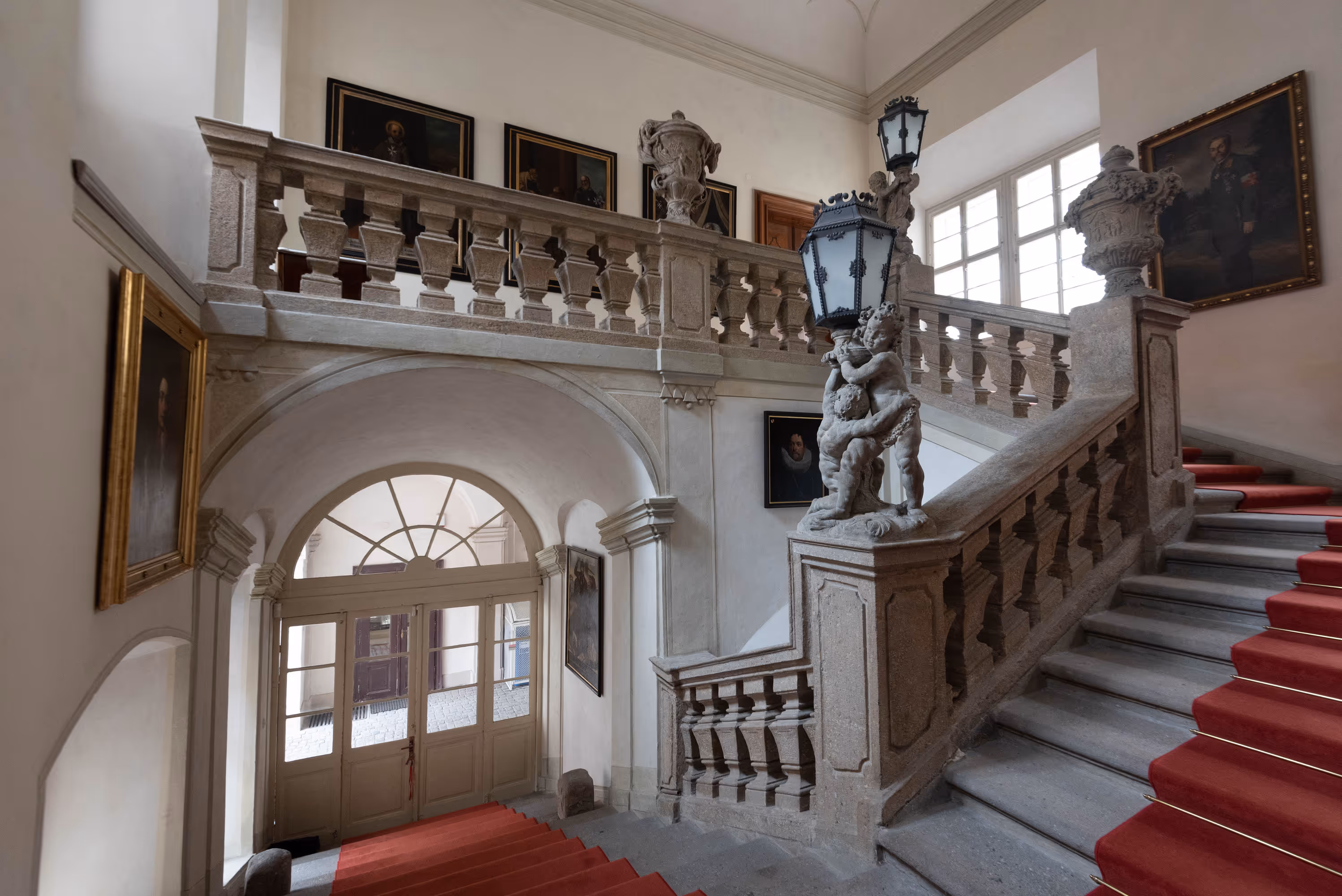 Ornate stone staircase inside a historic building with red carpet runners, decorative lamp posts featuring cherubs, and framed portraits on the walls.