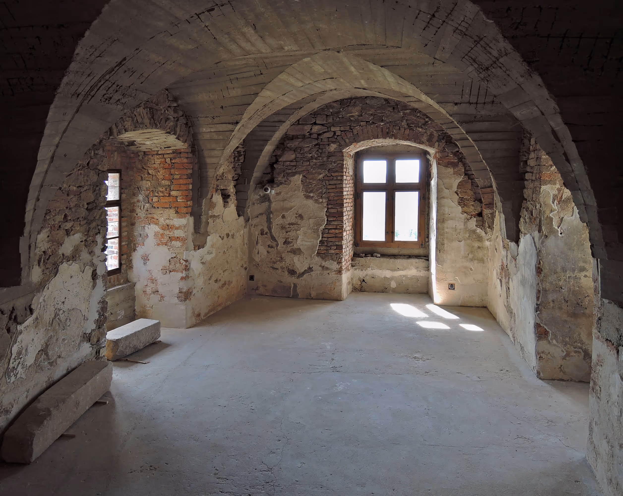 Empty room with vaulted ceilings, exposed brick and stone walls, and light coming through a tall window.
