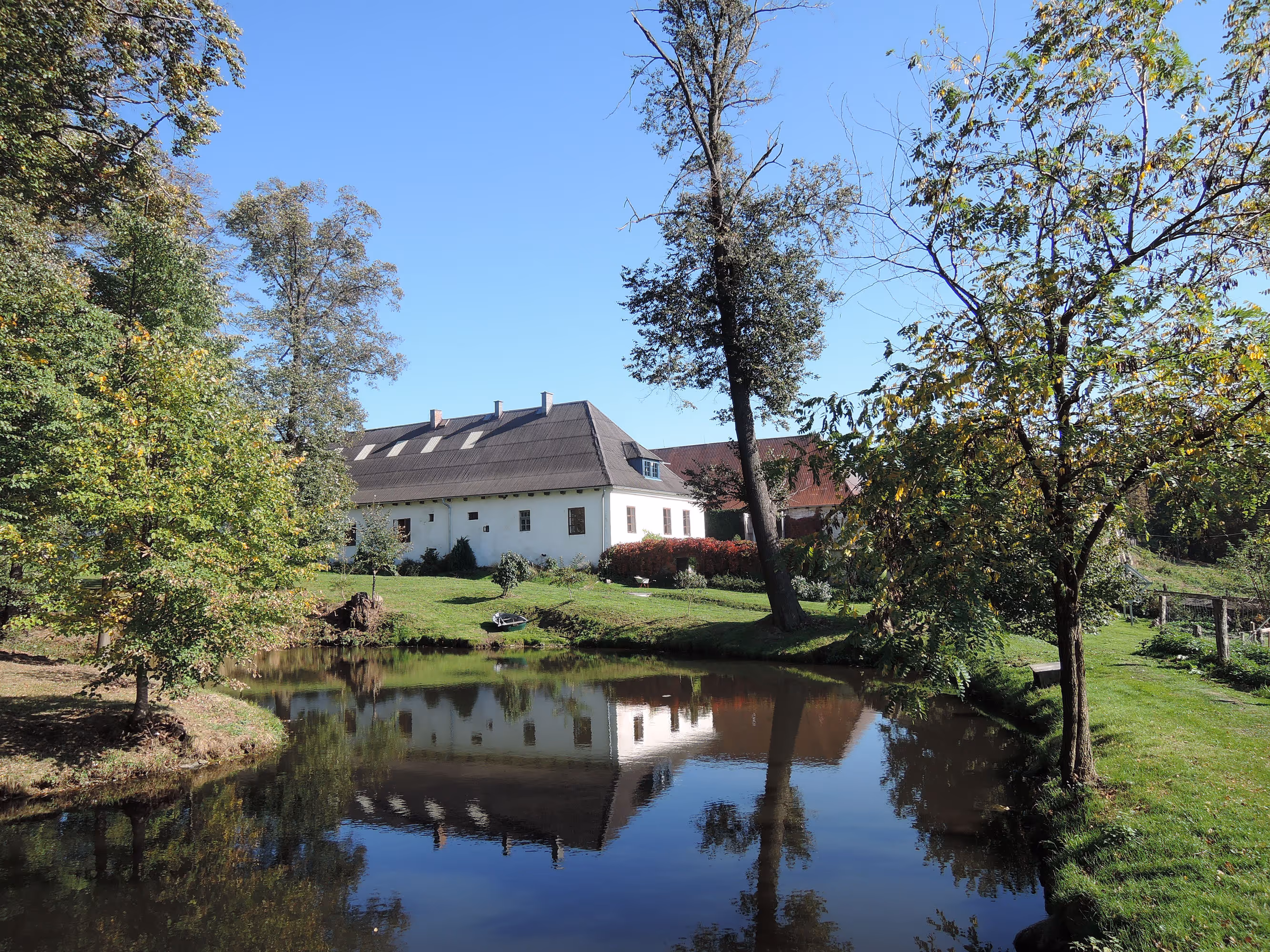 White house with a pitched roof next to a small pond reflecting the house and surrounding trees under a clear blue sky.