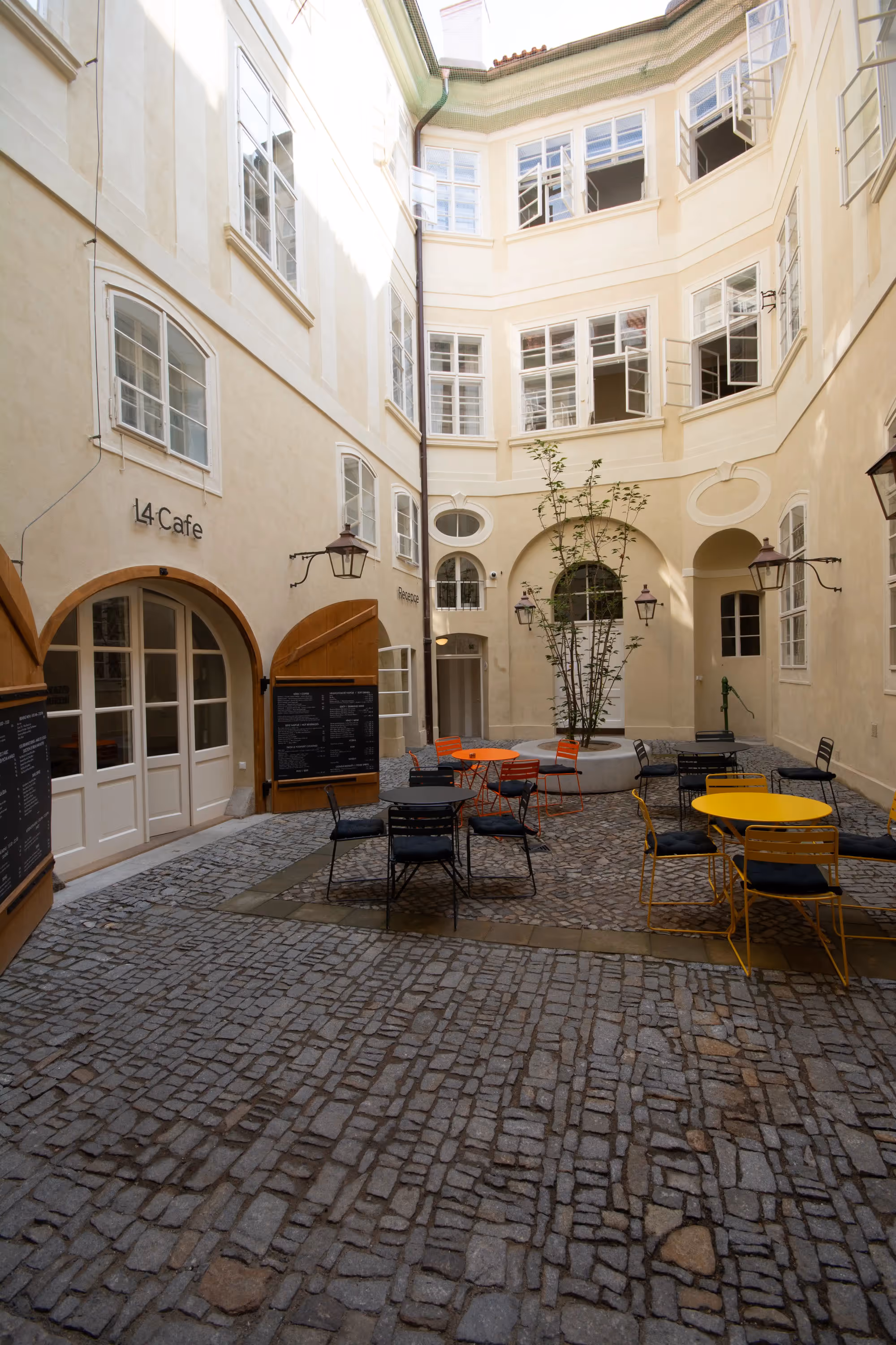 Sunny courtyard with cobblestone floor, colorful outdoor café tables and chairs, and cream-colored buildings with open windows.