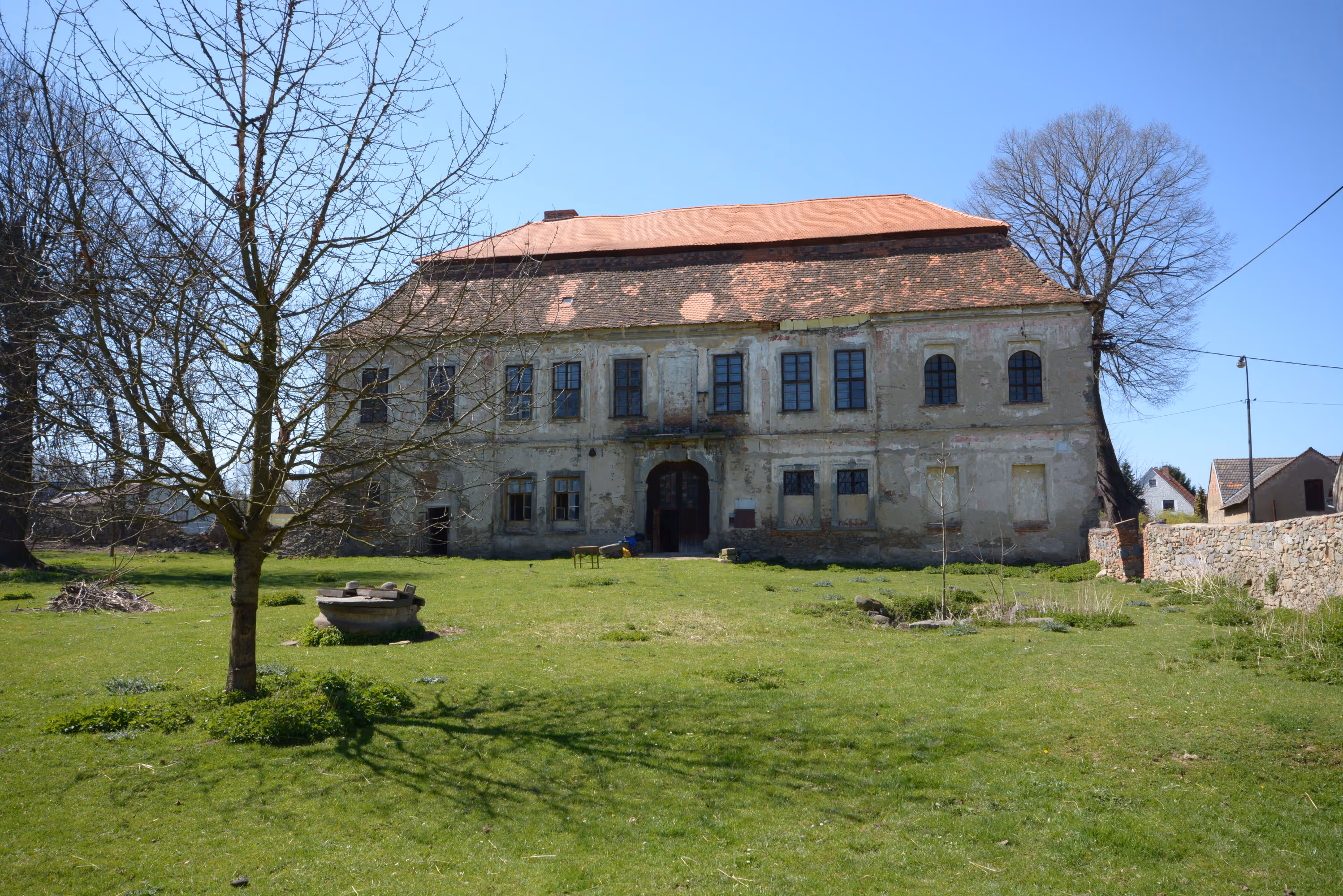 Old two-story building with worn facade and tiled roof, surrounded by green grass and leafless trees under a clear blue sky.
