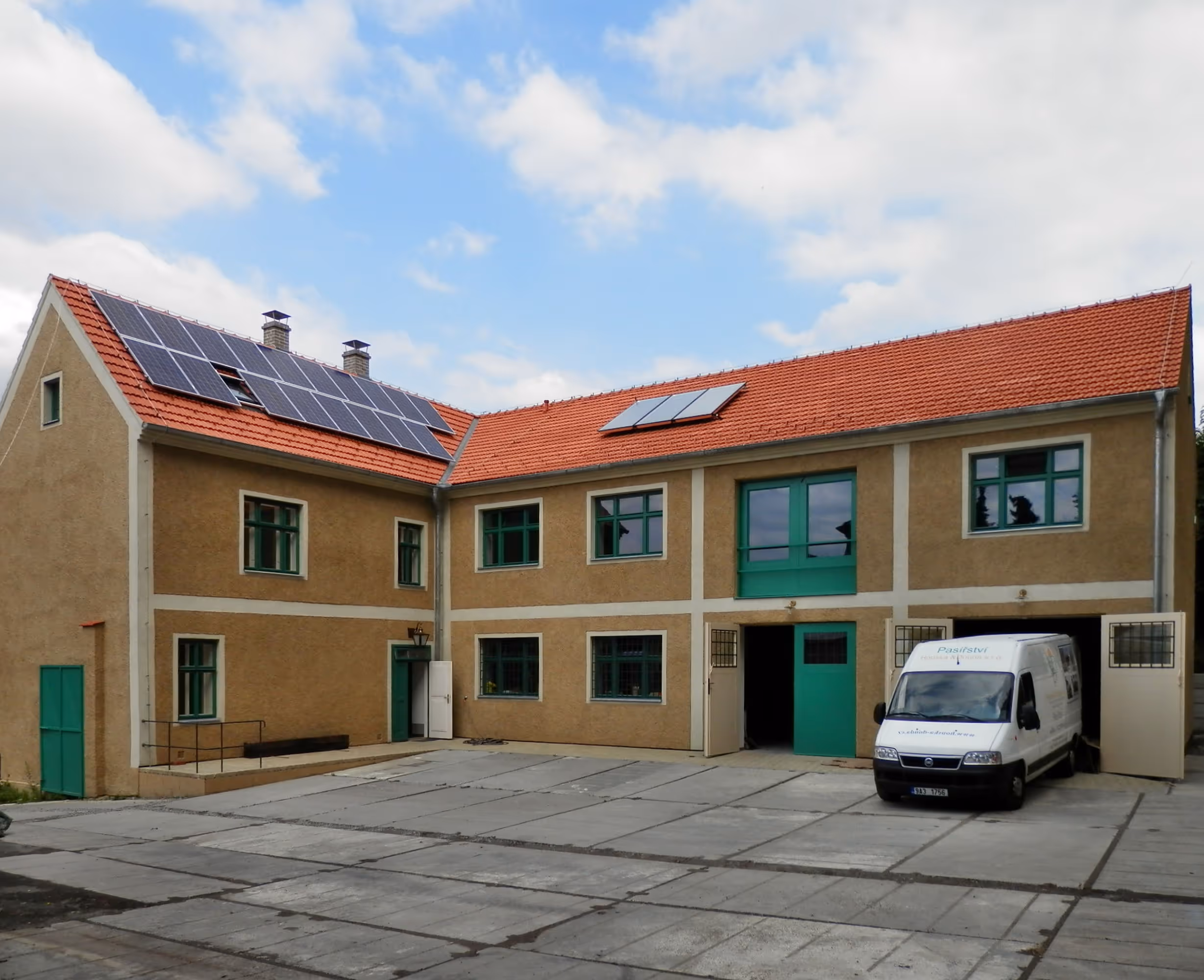 Two-story beige building with green windows and doors, solar panels on red-tiled roof, and a white van parked outside.