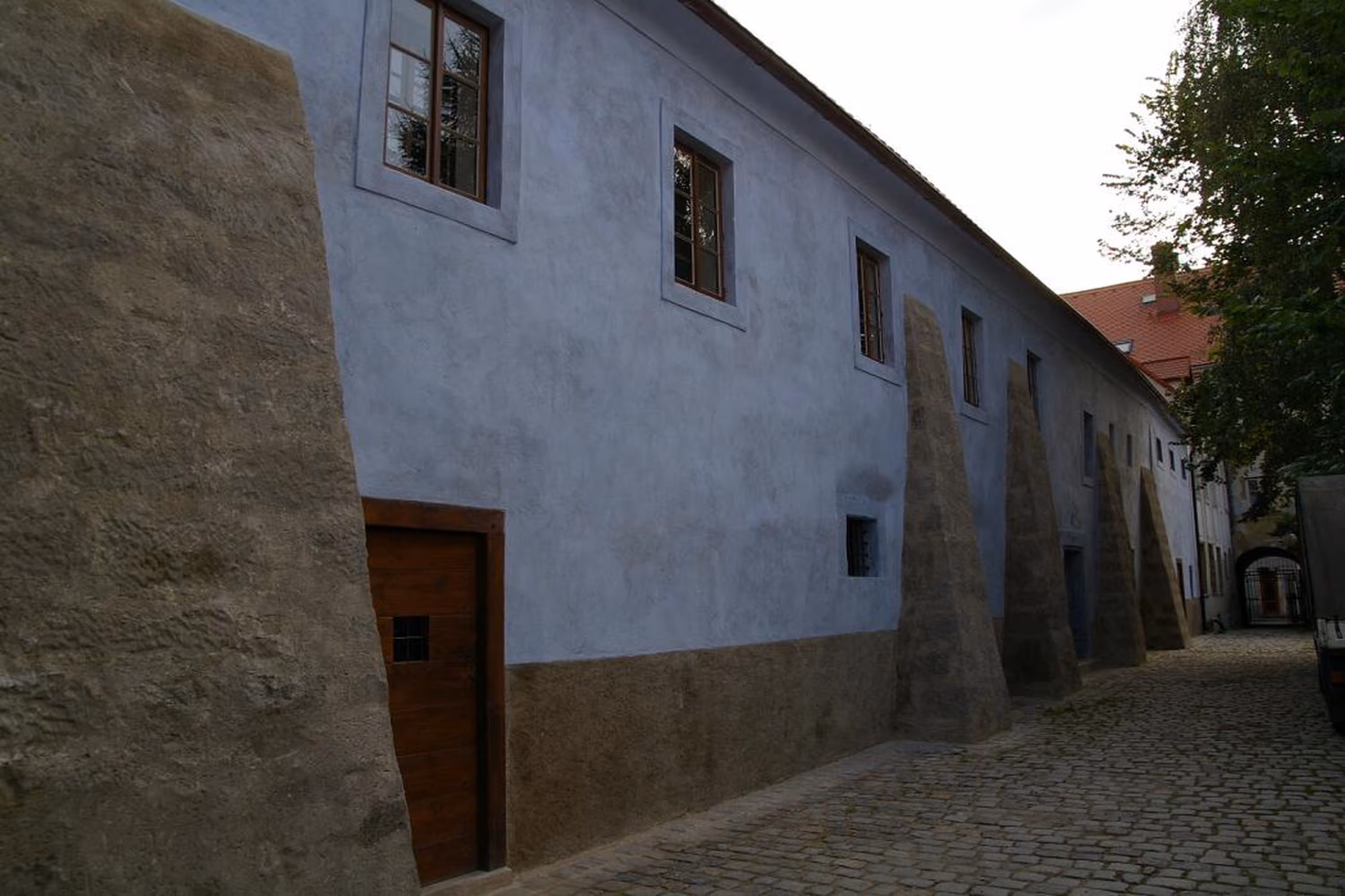 Long, pale blue building with triangular stone buttresses along a cobblestone street under an overcast sky.