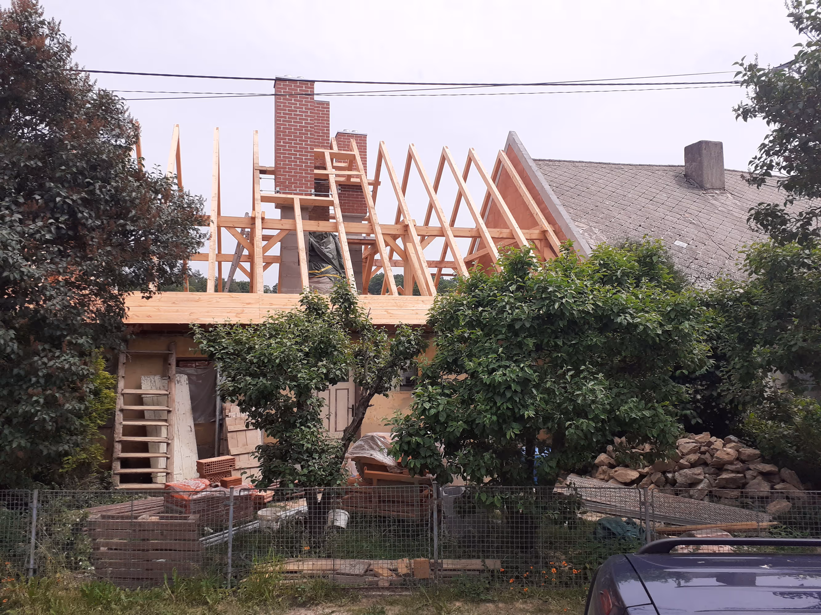 Partially constructed wooden roof frame with exposed beams and a red brick chimney, surrounded by green trees and garden materials.