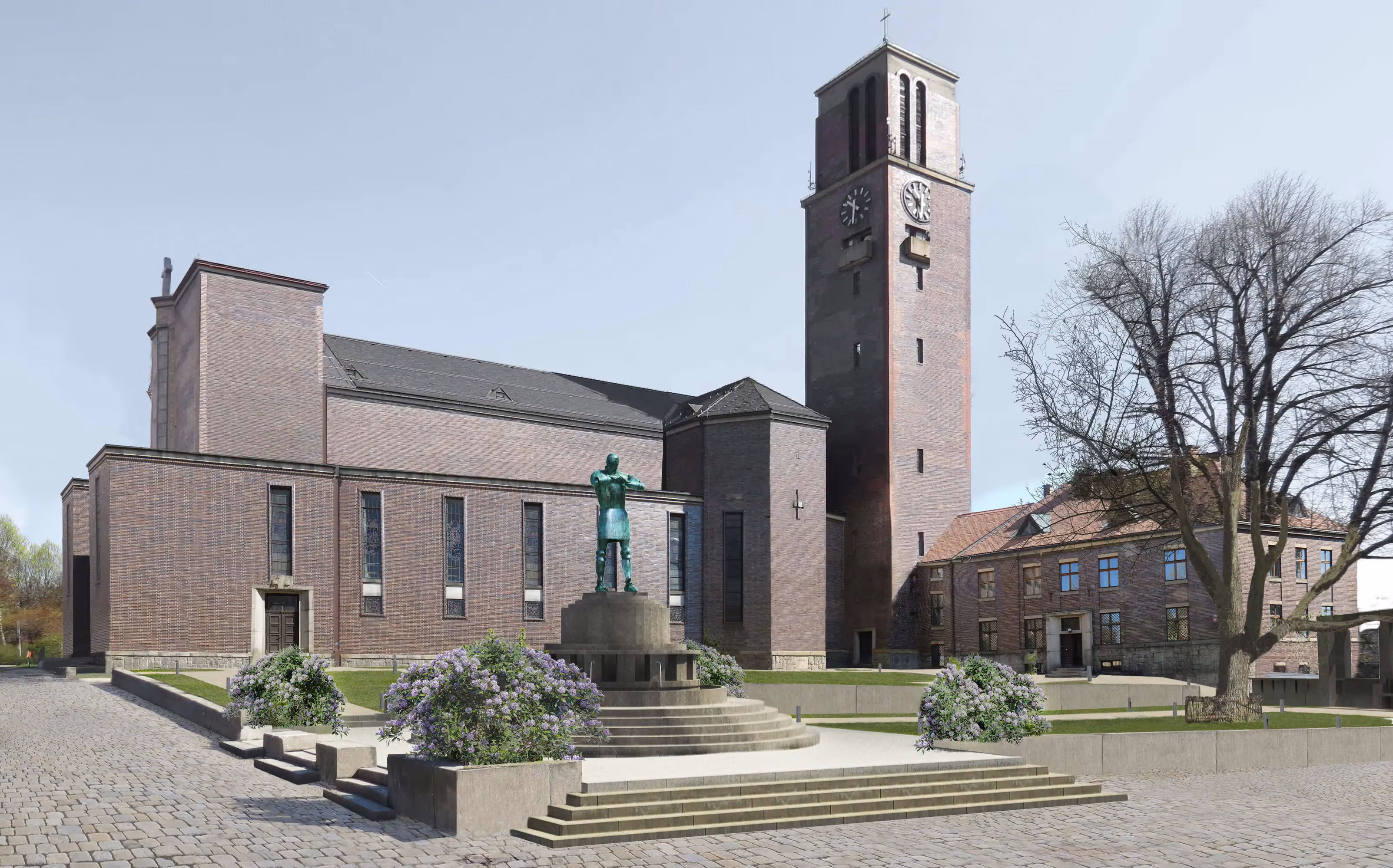 Brick church with a tall clock tower, a statue on a stepped pedestal, flowering bushes, and a leafless tree in front.