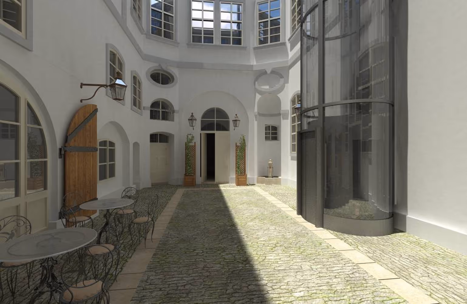 Sunlit cobblestone courtyard with round metal tables and chairs on the left and a modern glass elevator on the right.