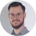 Porträt eines Mannes mit Brille, kurzem dunklem Haar und Bart, der lächelnd in die Kamera blickt.