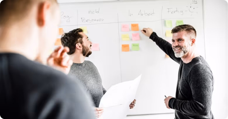 Drei Männer arbeiten gemeinsam an einem Whiteboard mit bunten Haftnotizen in einem Besprechungsraum.