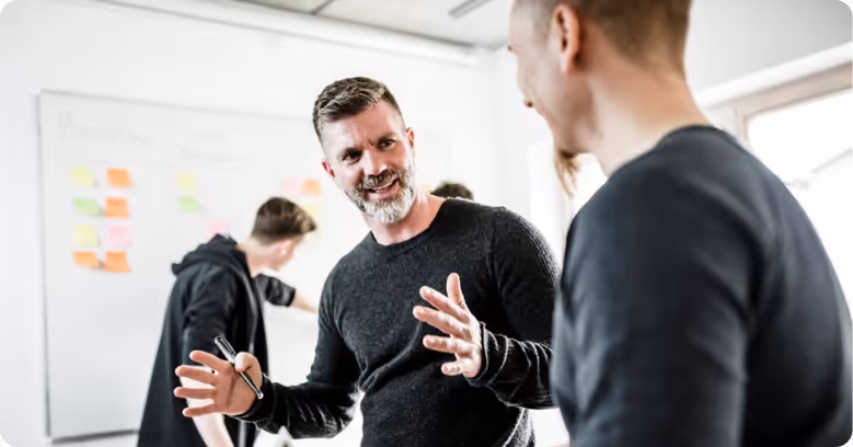 Zwei Männer im Gespräch vor einem Whiteboard mit bunten Haftnotizen, ein Mann gestikuliert mit den Händen.