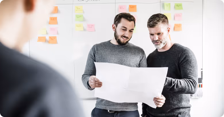 Zwei Männer betrachten gemeinsam ein großes Blatt Papier vor einer Wand mit bunten Haftnotizen.