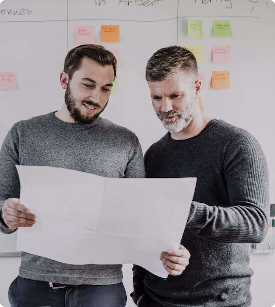 Zwei Männer lesen gemeinsam ein großes Blatt Papier vor einer Wand mit farbigen Haftnotizen.