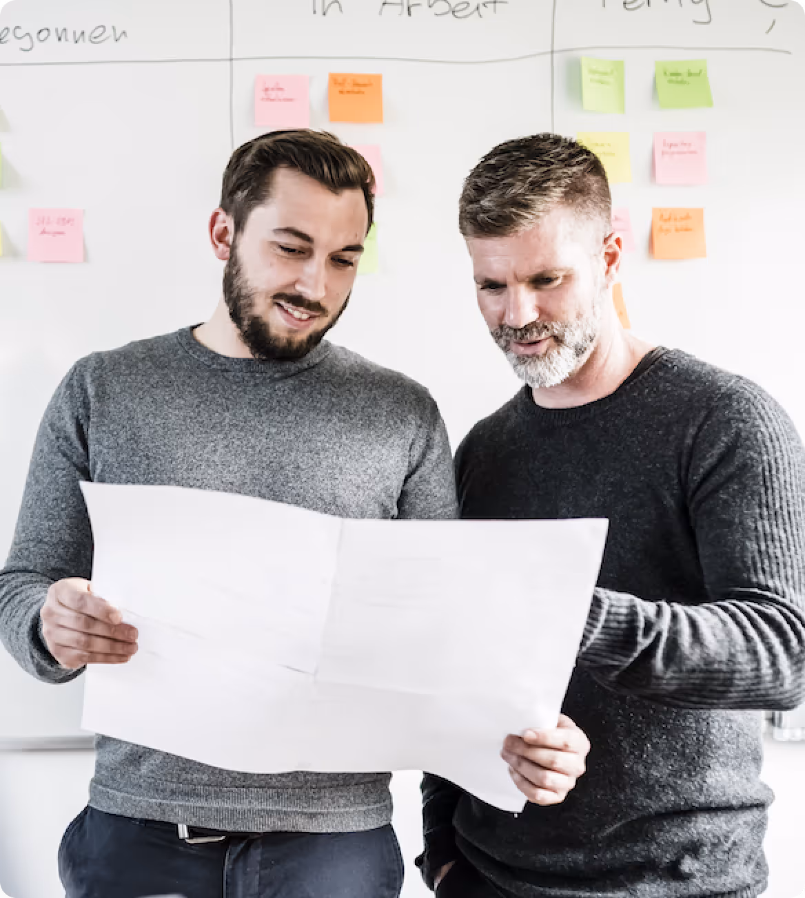 Zwei Männer in grauen Pullovern betrachten gemeinsam ein großes Blatt Papier vor einer Wand mit bunten Haftnotizen.