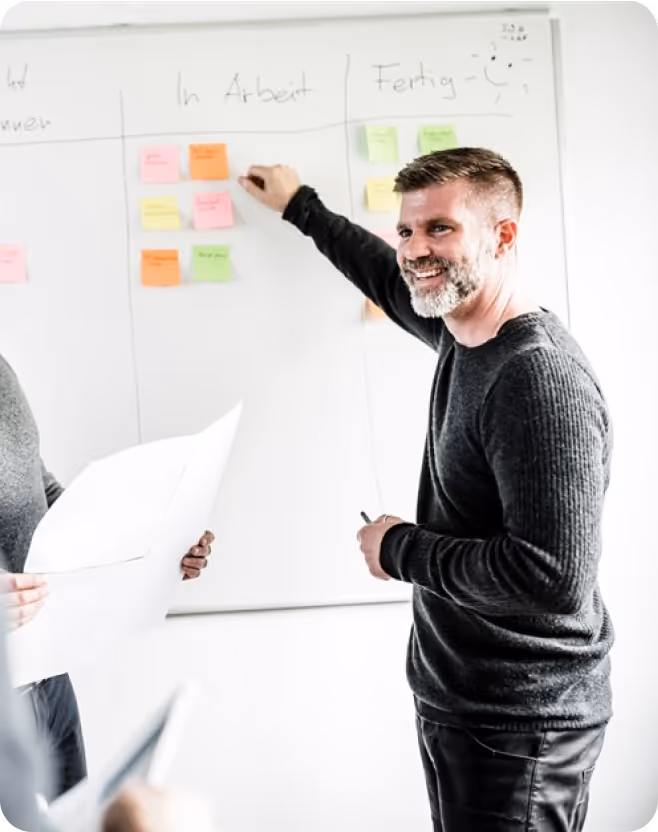 Mann lächelt und platziert bunte Haftnotizen auf einem Whiteboard mit den Überschriften 'In Arbeit' und 'Fertig'.