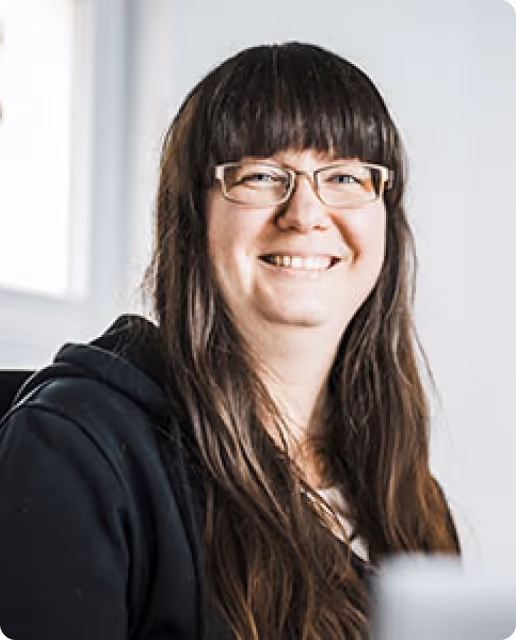 Porträt einer lächelnden Frau mit langen braunen Haaren und Brille, die einen schwarzen Hoodie trägt.