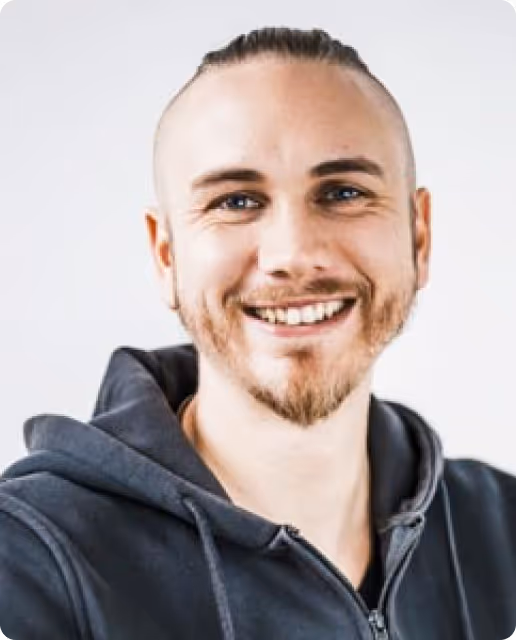 Junger Mann mit blauen Augen, kurzem Zopf, Bart und Kapuzenjacke, der lächelt.