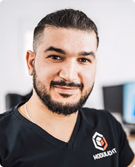 Porträt eines lächelnden Mannes mit kurzem dunklem Haar und Bart, der ein schwarzes Hemd mit dem Logo von Modulbot trägt.