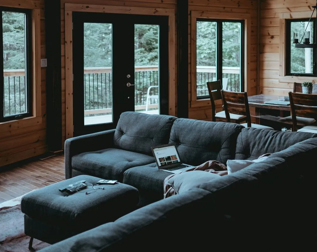 Cozy wooden cabin interior featuring a dark gray sofa, wooden dining table, and large windows with views of greenery outside.