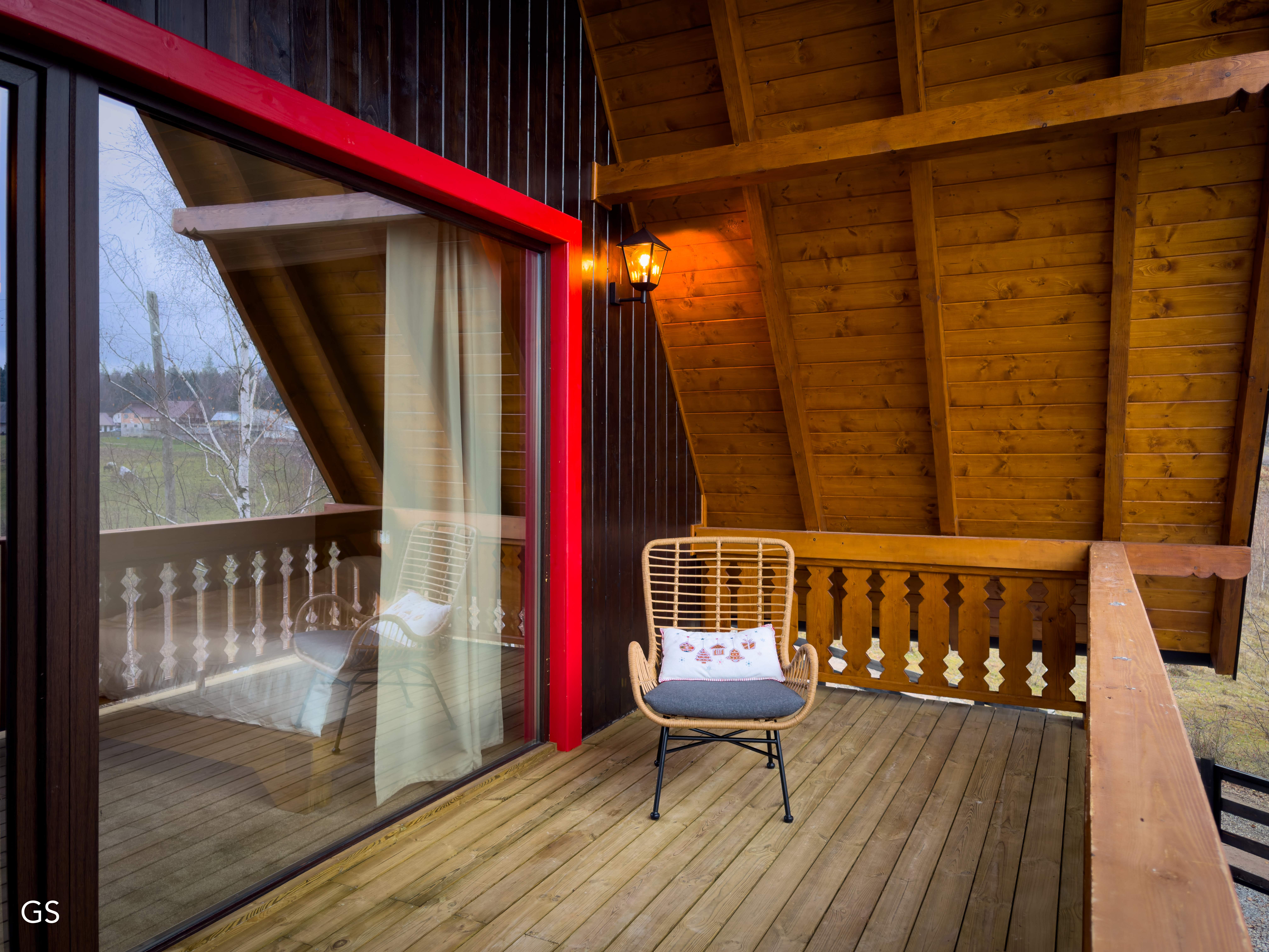 Cozy, modern living room with angled wood beams, large windows, a light gray sofa, and a decorative glass coffee table. Greenery visible outdoors.