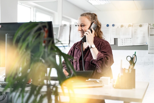 Smiling woman talking on a desk phone in a bright office with papers pinned on the wall behind her.
