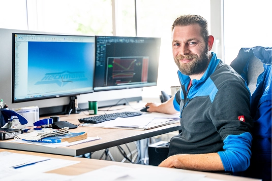 Man with beard and blue-gray jacket sitting at a desk with dual computer monitors displaying design software.