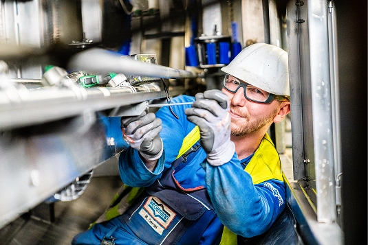 Worker in blue and yellow safety uniform and helmet repairing or inspecting metal machinery components.