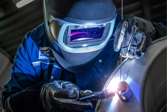 Welder in protective gear using a welding torch on metal in a dimly lit workshop.