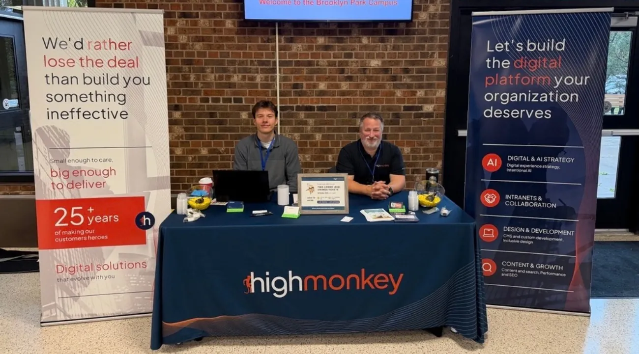 The High Monkey M365 Twin Cities booth with two banners on either side of a table, all containing High Monkey branding. Cole and Chad sitting at the table smiling.