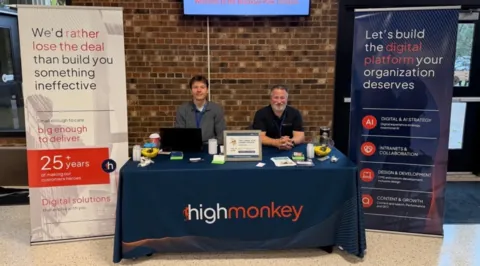 The High Monkey M365 Twin Cities booth with two banners on either side of a table, all containing High Monkey branding. Cole and Chad sitting at the table smiling.