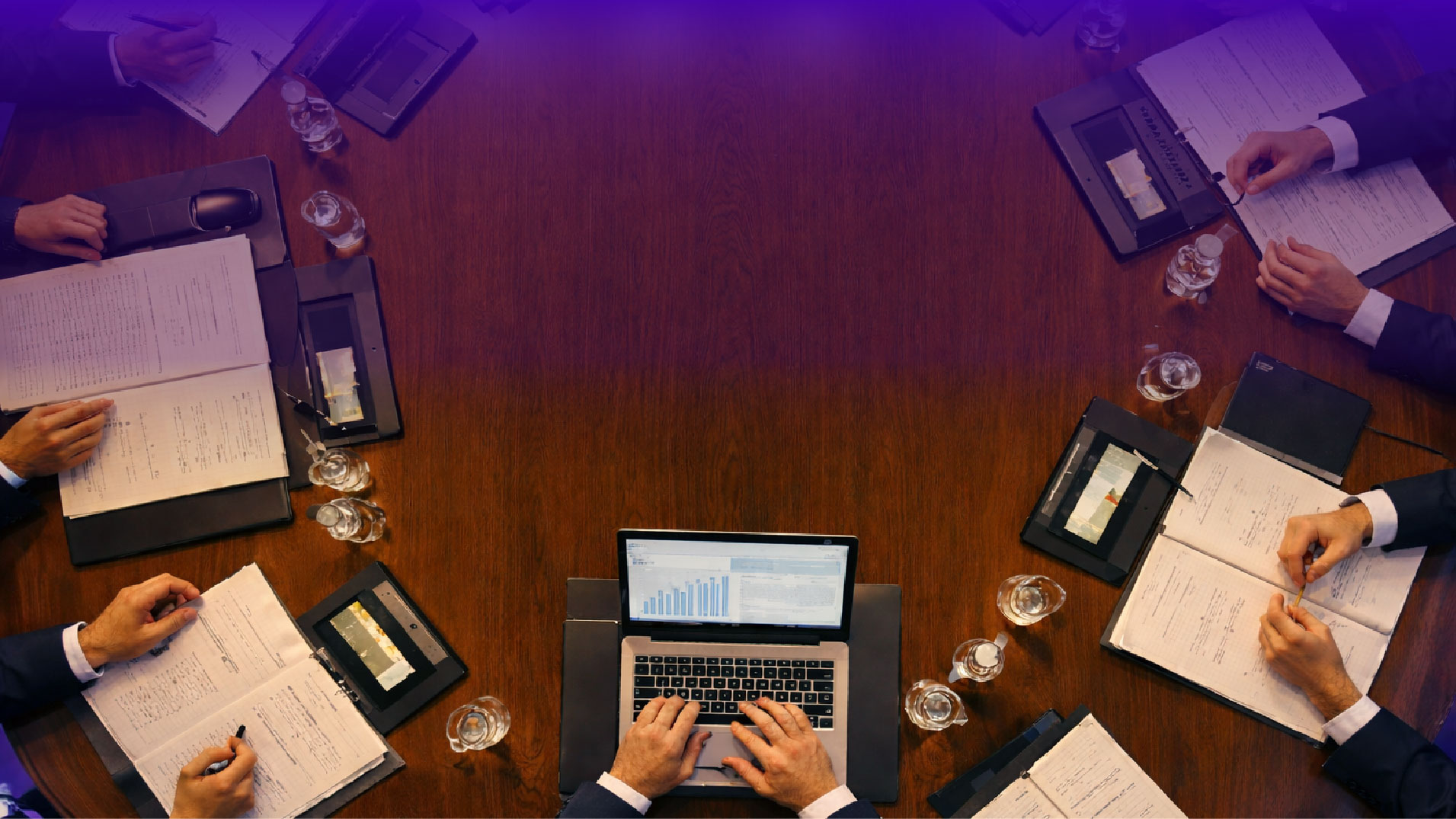 Vista dall’alto di un tavolo da riunione in legno con professionisti seduti attorno, documenti aperti, tablet, bicchieri d’acqua e un laptop al centro con grafici sullo schermo