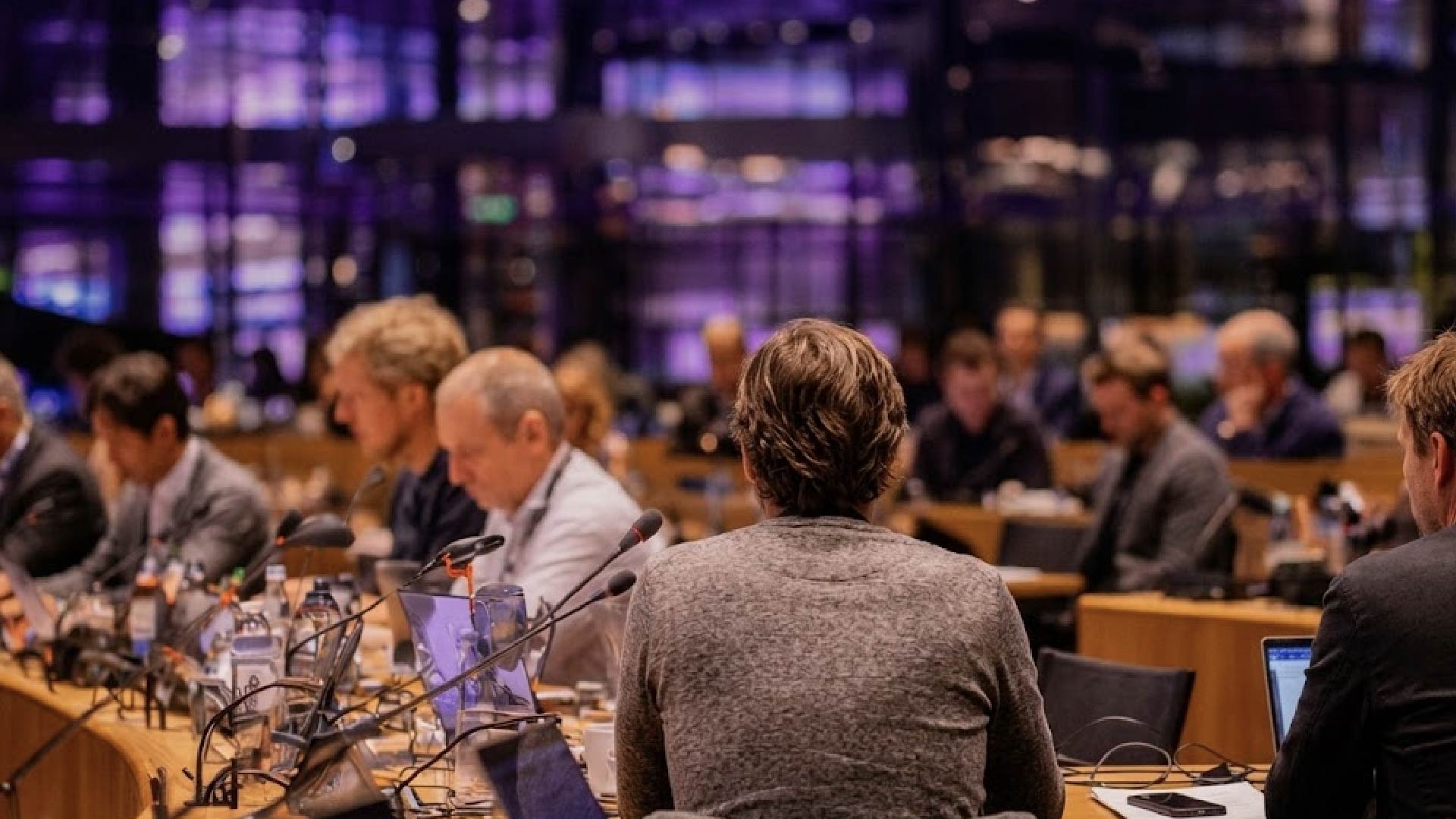 Persona di spalle seduta a un tavolo durante una conferenza, circondata da altri partecipanti con microfoni