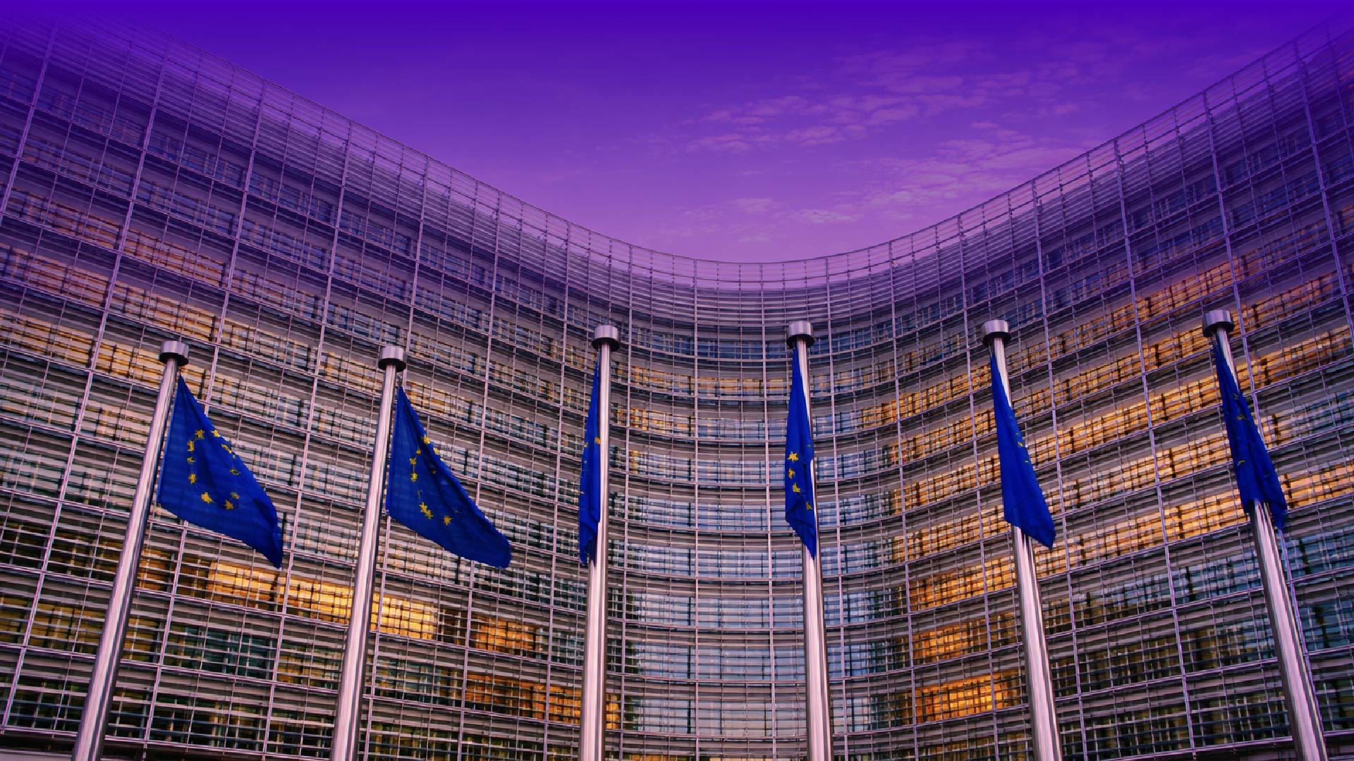 Facciata curva di un moderno edificio istituzionale in vetro (sede dell’Unione Europea) con diverse bandiere blu dell’UE su alti pennoni in primo piano