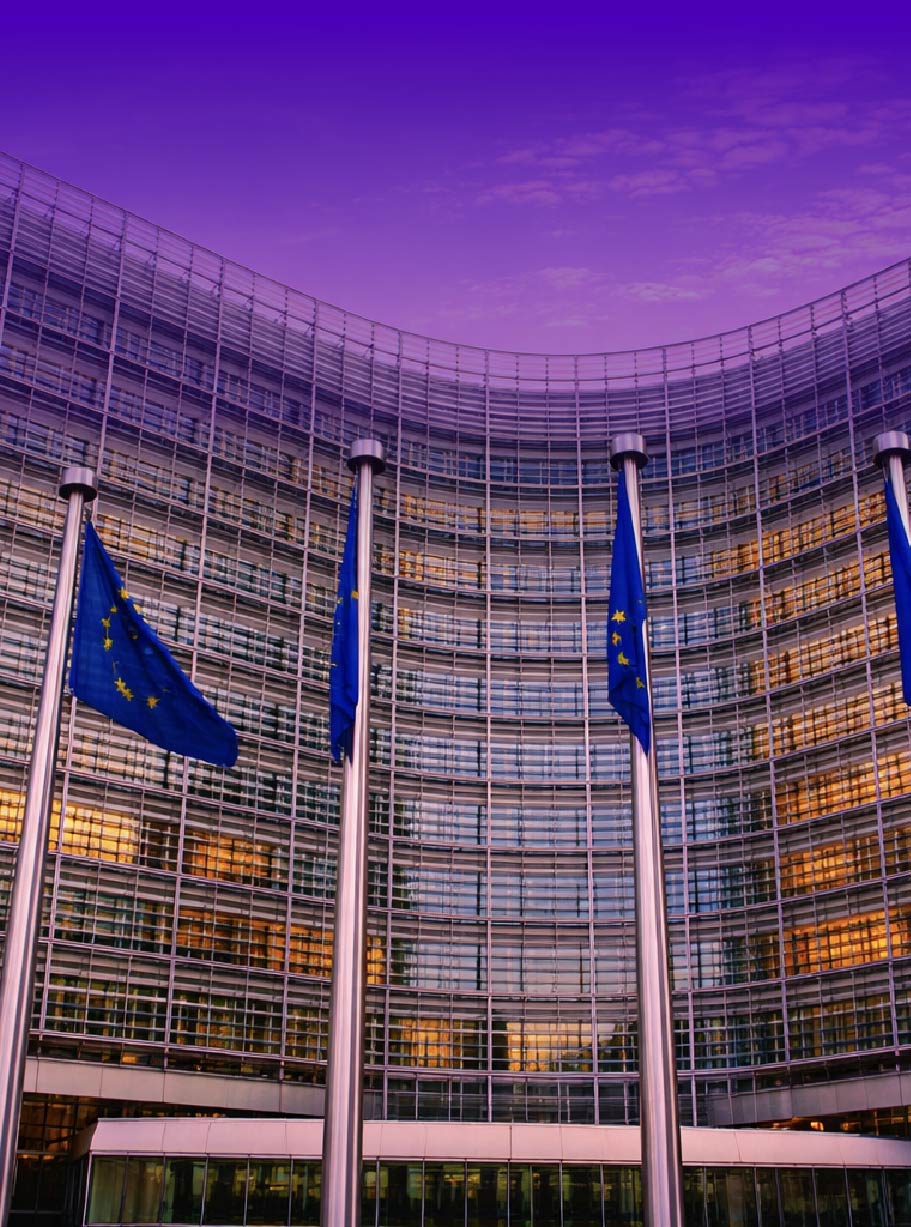 Curved glass facade of a modern institutional building (European Union headquarters) with several blue EU flags on tall flagpoles in the foreground