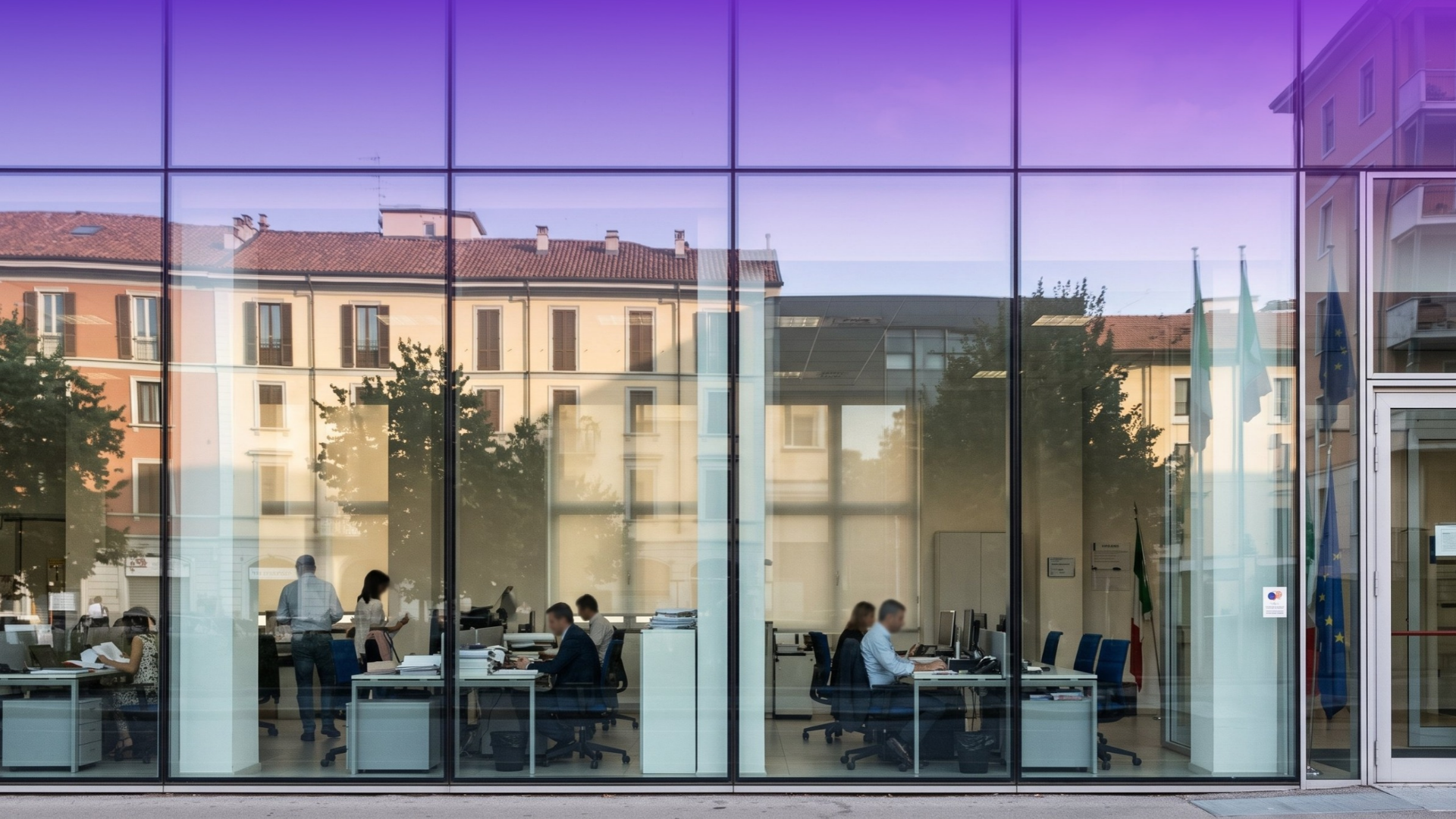 Persone al lavoro negli uffici di un edificio pubblico, visibili attraverso un'ampia facciata vetrata che riflette gli edifici circostanti