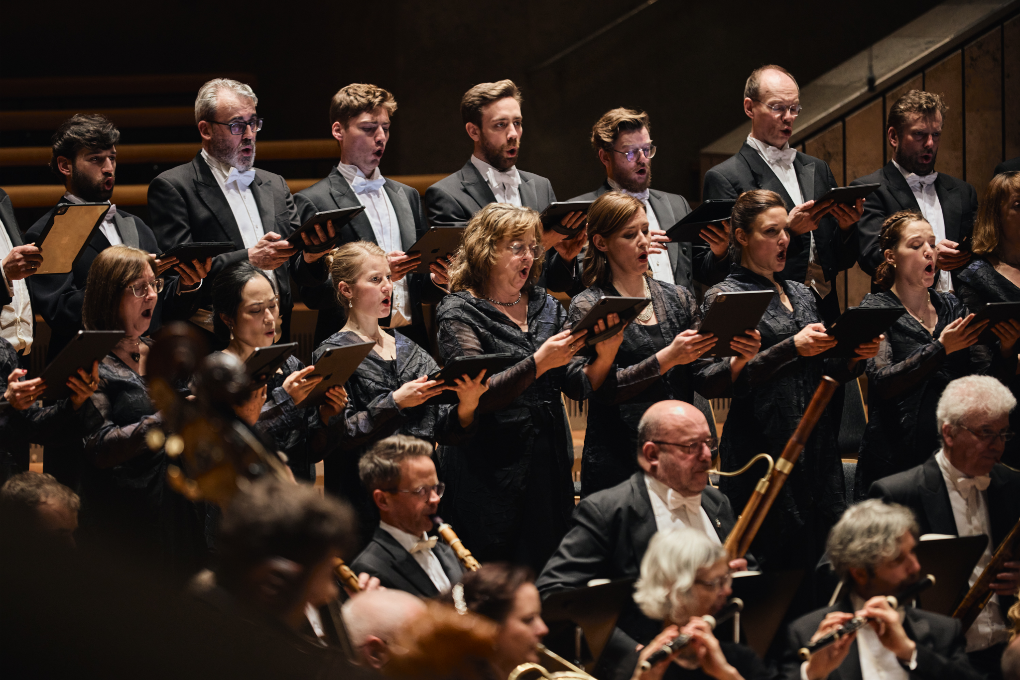 Un chœur d'hommes et de femmes en vêtements formels noirs chantant ensemble dans une salle de concert, tenant des tablettes pour lire la musique, avec des musiciens en bas jouant des instruments à vent.