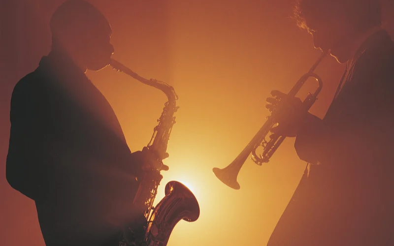 Silhouettes de deux musiciens jouant du saxophone et de la trompette devant un éclairage chaud orangé.