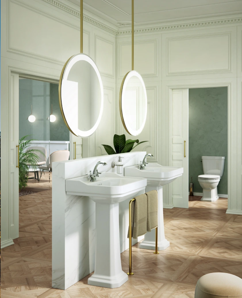 Radiance Visual. Elegant bathroom with two pedestal sinks separated by a marble divider, illuminated round mirrors, gold-toned towel rack with beige towels, and a toilet in an adjacent room.