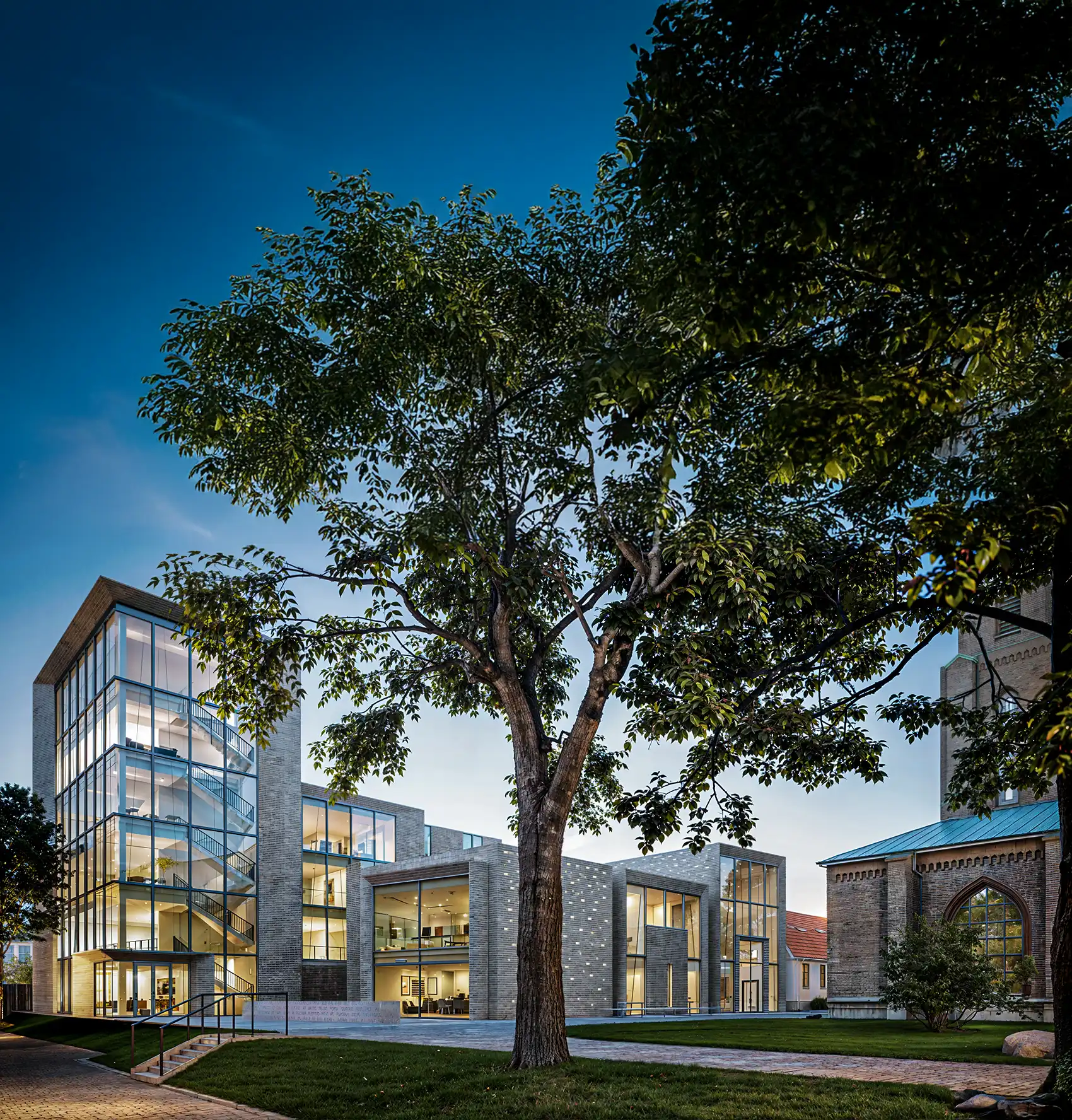 Radiance Visual. Modern building with large glass windows and visible staircases next to an older brick church, framed by trees and a blue sky at dusk.