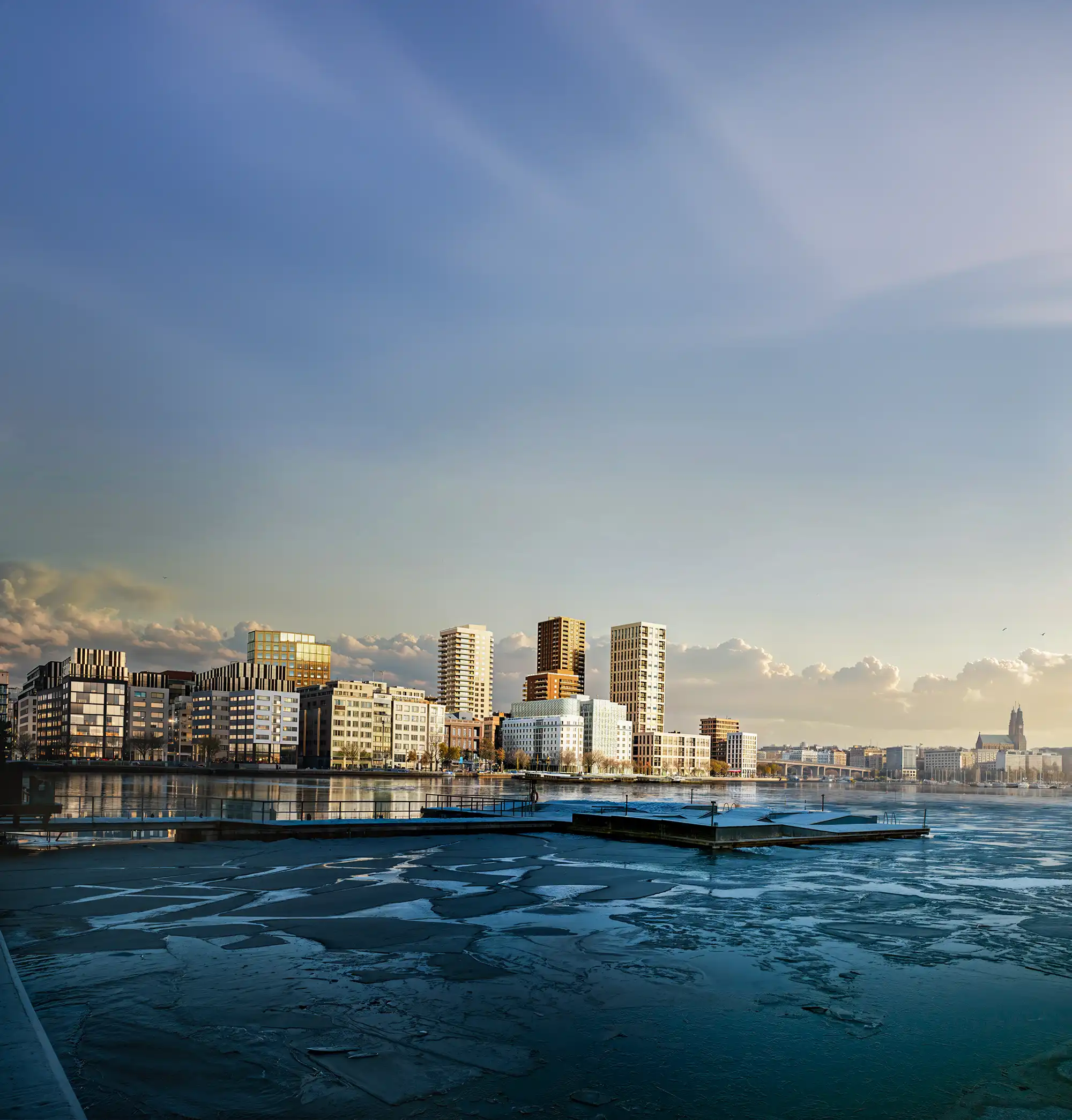 Radiance Visual. City skyline with modern buildings along a partially frozen waterfront under a clear sky with scattered clouds.