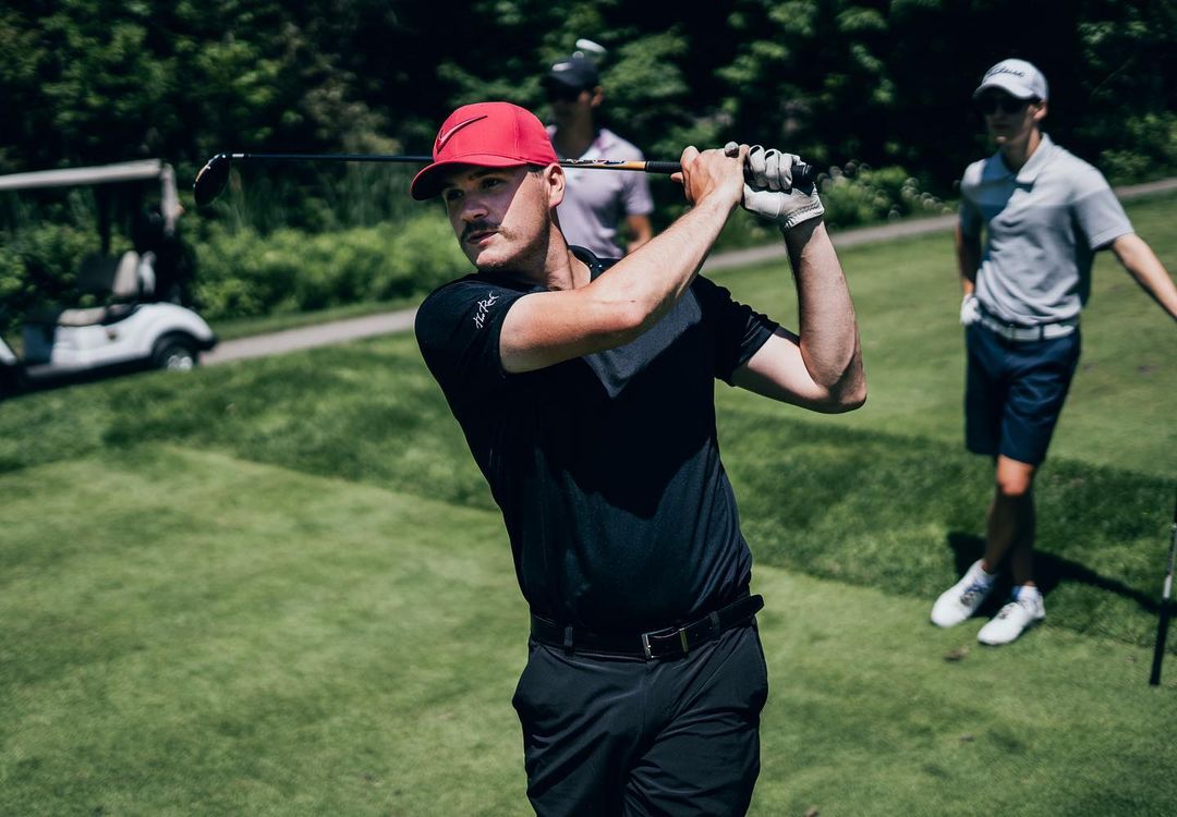 Golfer in a red hat finishing a swing on a sunny course with other players and a golf cart in the background.
