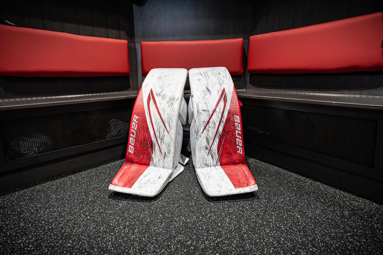 Pair of red and white Bauer goalie pads leaning against a bench inside a hockey locker room.