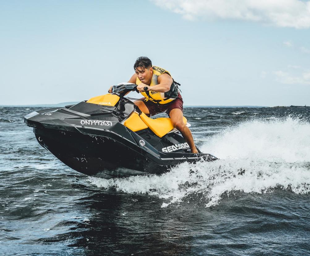 Person riding a yellow Sea-Doo jet ski at high speed over choppy water under a partly cloudy sky.