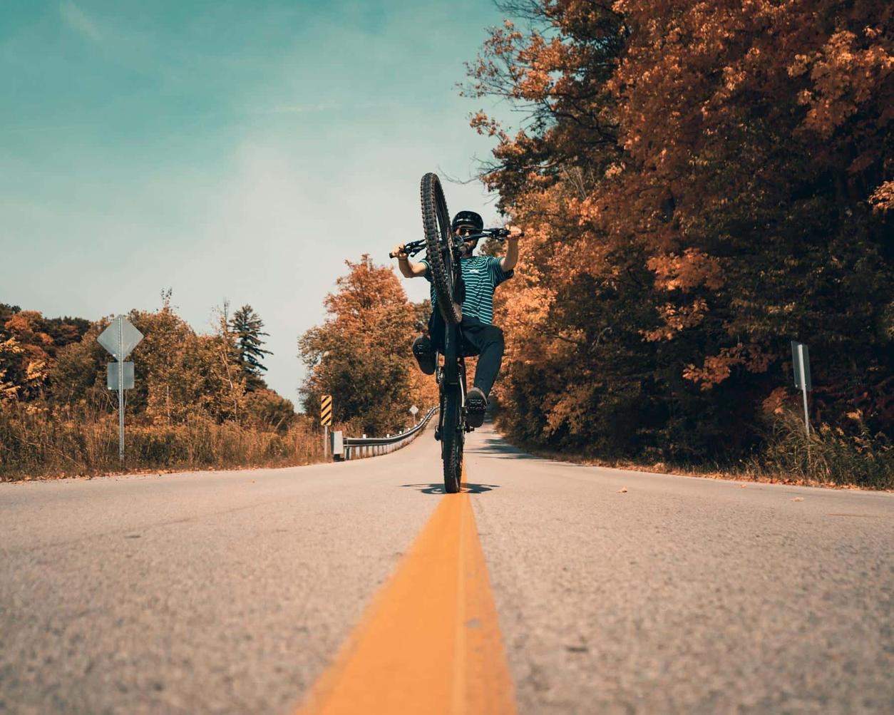 Cyclist performing a wheelie on a rural road lined with autumn trees under a clear blue sky.