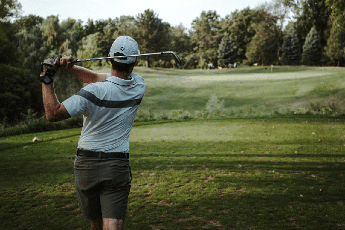 Golfer viewed from behind finishing a swing on a green fairway surrounded by trees.