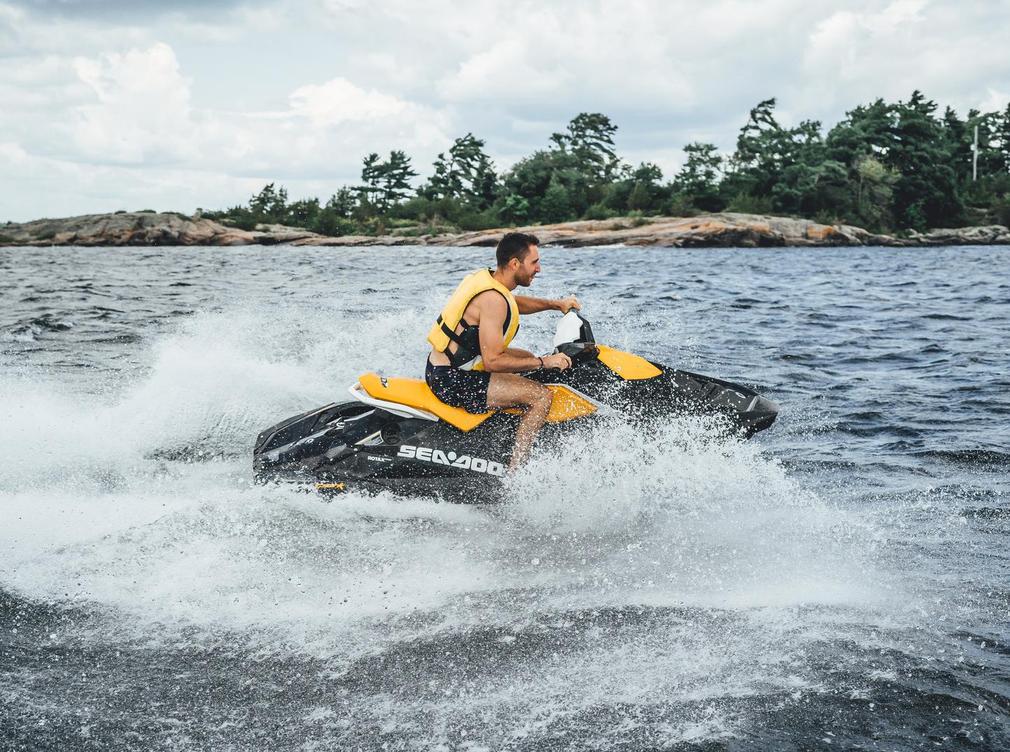 Person riding a yellow jet ski across a lake, creating splashes with trees and rocky shoreline in the background.