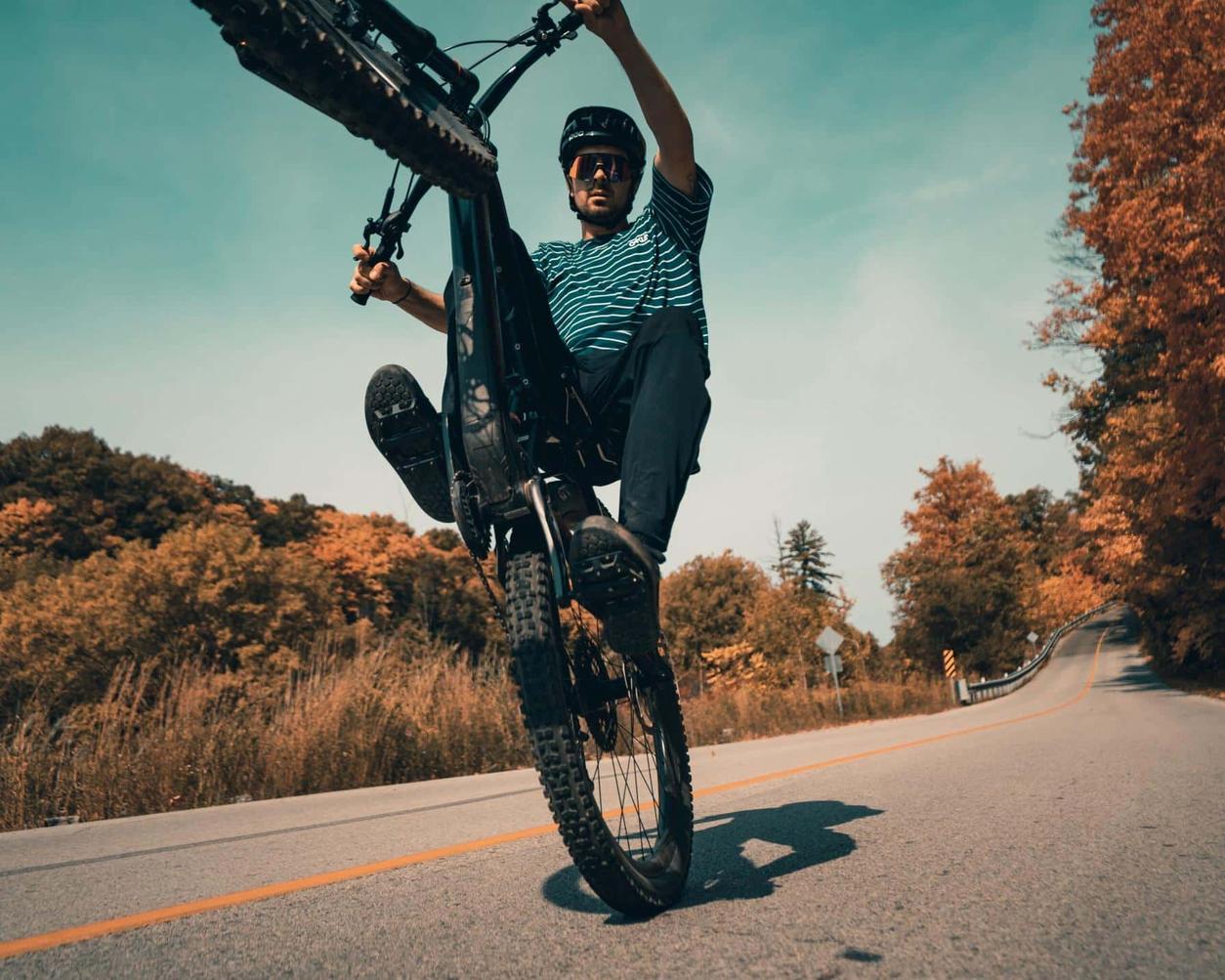 Mountain biker performing a wheelie on a scenic road surrounded by autumn trees under a clear sky.